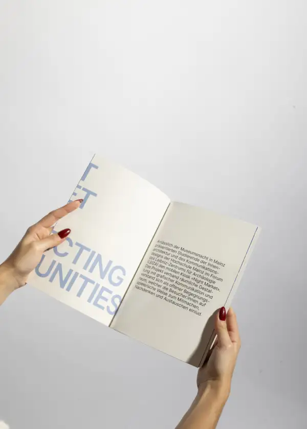 Hände mit roten Fingernägeln halten ein aufgeschlagenes Buch mit Text auf der rechten Seite und großem blauen Text auf der linken Seite vor hellem Hintergrund.