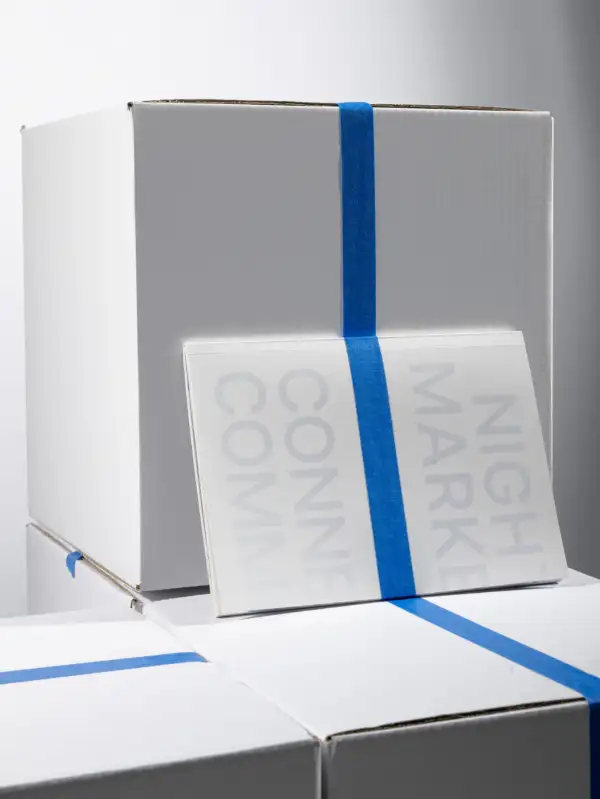 Drei weiße Kartons mit blauen Klebestreifen, auf einem Karton liegt ein weißes Softcover mit blassem Text 'NIGHT MARKET'