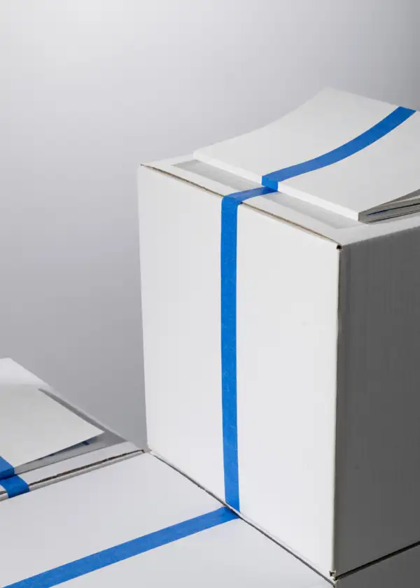 Drei weiße Kartons mit blauen Klebestreifen, auf einem Karton liegt ein weißes Softcover mit blassem Text 'NIGHT MARKET'