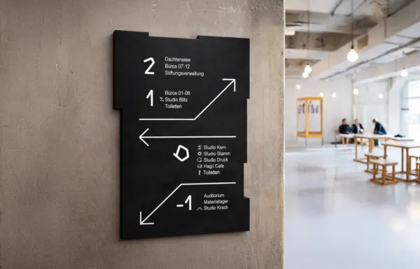 Schwarze Leittafel mit weißer Typografie an Betonwand, Angaben zu Stockwerken, rechts im Hintergrund Personengruppe an Holztischen.