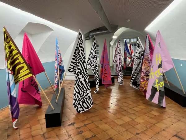 Bunte Fahnen mit Typografie auf Holzstäben, schwarze Holzblöcke auf dem Boden als Halterung, Brauner Holzboden