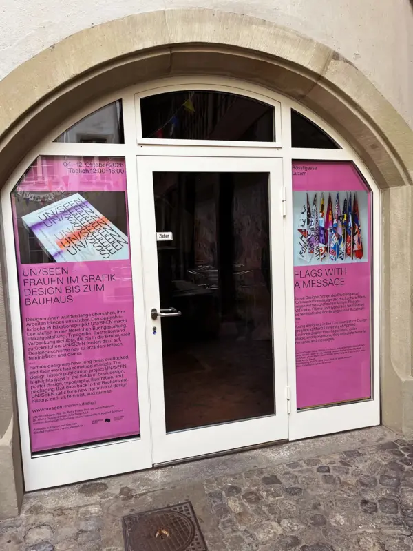 Weiß schwarze Eingangstür in Steinbogen, links und rechts zwei große Plakate in den Scheiben, Pink mit schwarzer Schrift und jeweils ein Bild,